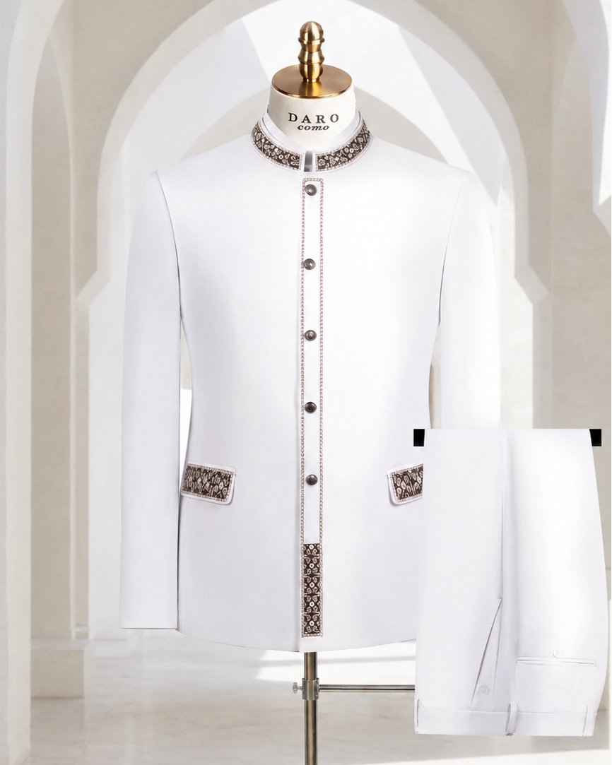 A white traditional men's jacket and matching trousers displayed on a mannequin stand in a bright, arched interior. The jacket has decorative patterned trim at the collar, front placket, and pocket flaps, with dark buttons.