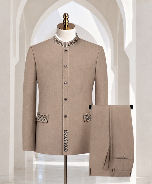 A beige men's traditional suit set consisting of a jacket displayed on a mannequin and matching trousers hanging beside it, set against an arched white marble interior background.