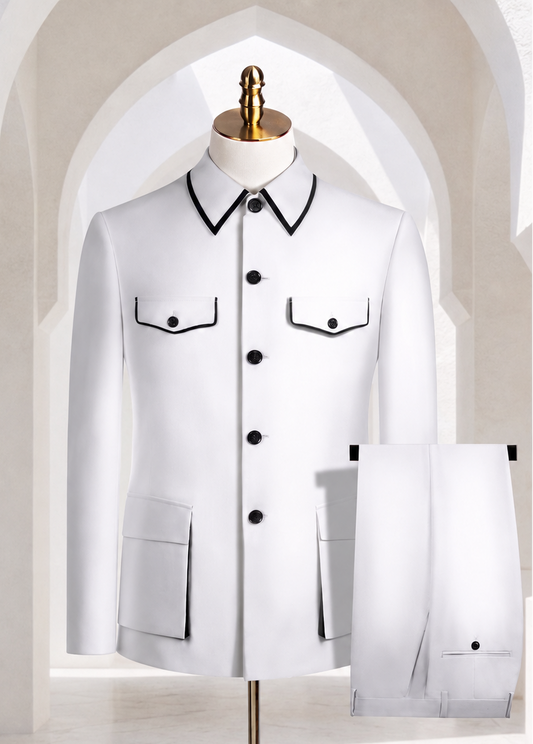 A white men's suit jacket and matching trousers displayed on a mannequin stand in a bright, arched interior. The jacket has black piping trim around the pointed collar, button placket, chest flap pockets, and front pockets, with matching black buttons.