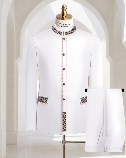 A white traditional men's jacket and matching trousers displayed on a mannequin stand in a bright, arched interior. The jacket has decorative patterned trim at the collar, front placket, and pocket flaps, with dark buttons.
